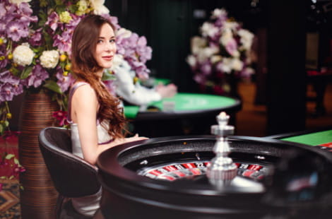 Long-haired redhead croupier in a gambling hall, sitting sideways next to a roulette wheel, sideways, with a standing vase with flowers and another gambling table in the background.