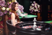 Long-haired redhead croupier in a gambling hall, sitting sideways next to a roulette wheel, sideways, with a standing vase with flowers and another gambling table in the background.