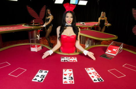 Playboy blackjack tables with four tables. In the foreground, a blackjack hand appears with a brunette dealer presenting the table with her palms facing up and dressed in a red bodysuit, bow tie and bunny ears. In the background there are two other Playboy bunnies leaning on two other casino tables.