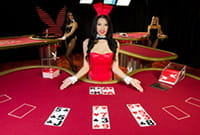 Playboy blackjack tables with four tables. In the foreground, a blackjack hand appears with a brunette dealer presenting the table with her palms facing up and dressed in a red bodysuit, bow tie and bunny ears. In the background there are two other Playboy bunnies leaning on two other casino tables.