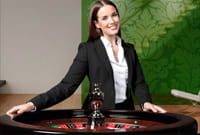 Smiling croupier dressed in a suit jacket and a white shirt with her arms resting on a roulette wheel, in a room with a green background.