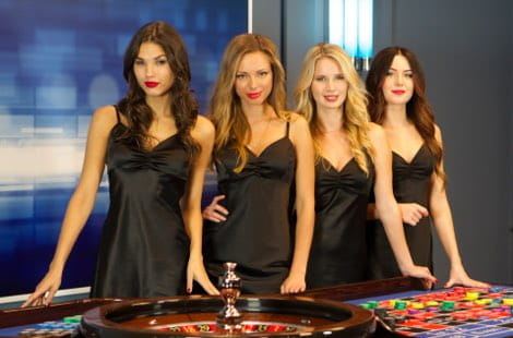 Four female croupiers dressed in similar black dresses, standing at a table with a roulette wheel and a doily, in a studio with a blue and white background.