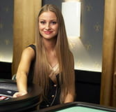 A croupier dressed in a black vest and sitting at a roulette table with her hand on the cylinder as if she were going to spin it.