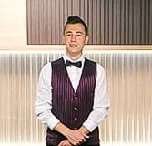 A dealer dressed in a white shirt, purple vest and black bow tie talking to the camera with his hands clasped at the level of the abdomen.