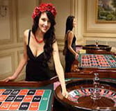 A croupier in a black strapless dress and a crown of roses on her head. She is leaning slightly on a casino table, looking at the camera and has her left arm on the cylinder as if she were going to spin it. In the background you can see another dealer at a different table.