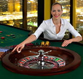 A croupier sitting at a casino table. He looks and smiles at the camera, and he has his right arm on the roulette cylinder as if he were going to spin it.